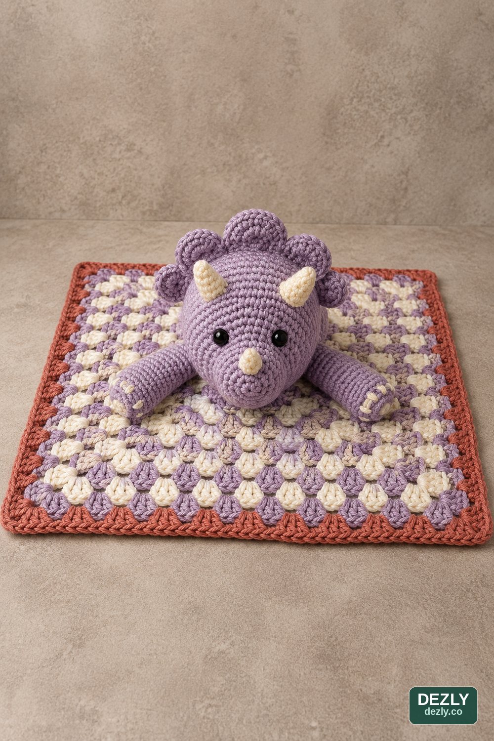 Finished crochet triceratops snuggle with a centered head and two legs on a granny-square blanket.